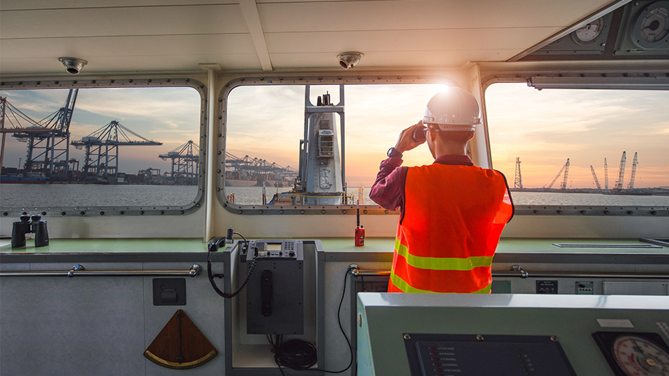 Man in safety vest and hard hat looking through binoculars from the bridge of a ship approaching port