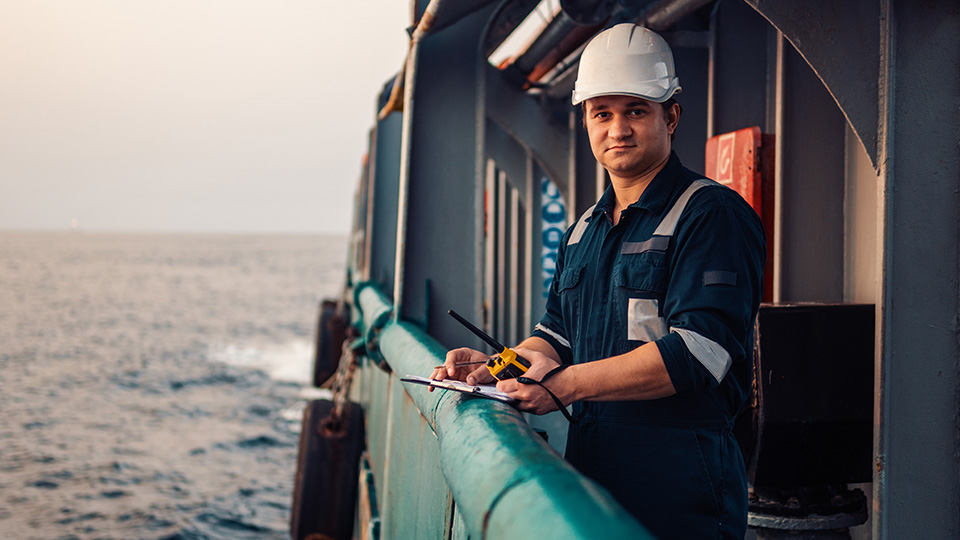 Deck officer standing at a ship's railing with a clipboard and walkie talkie.