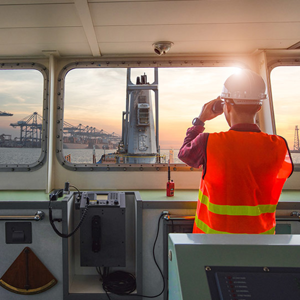 Man in safety vest and hard hat looking through binoculars from the bridge of a ship approaching port