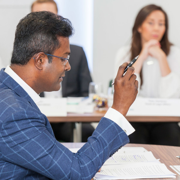 Man in suit sitting at a table reading papers and holding a pen up with a row of blurred people watching him in the background