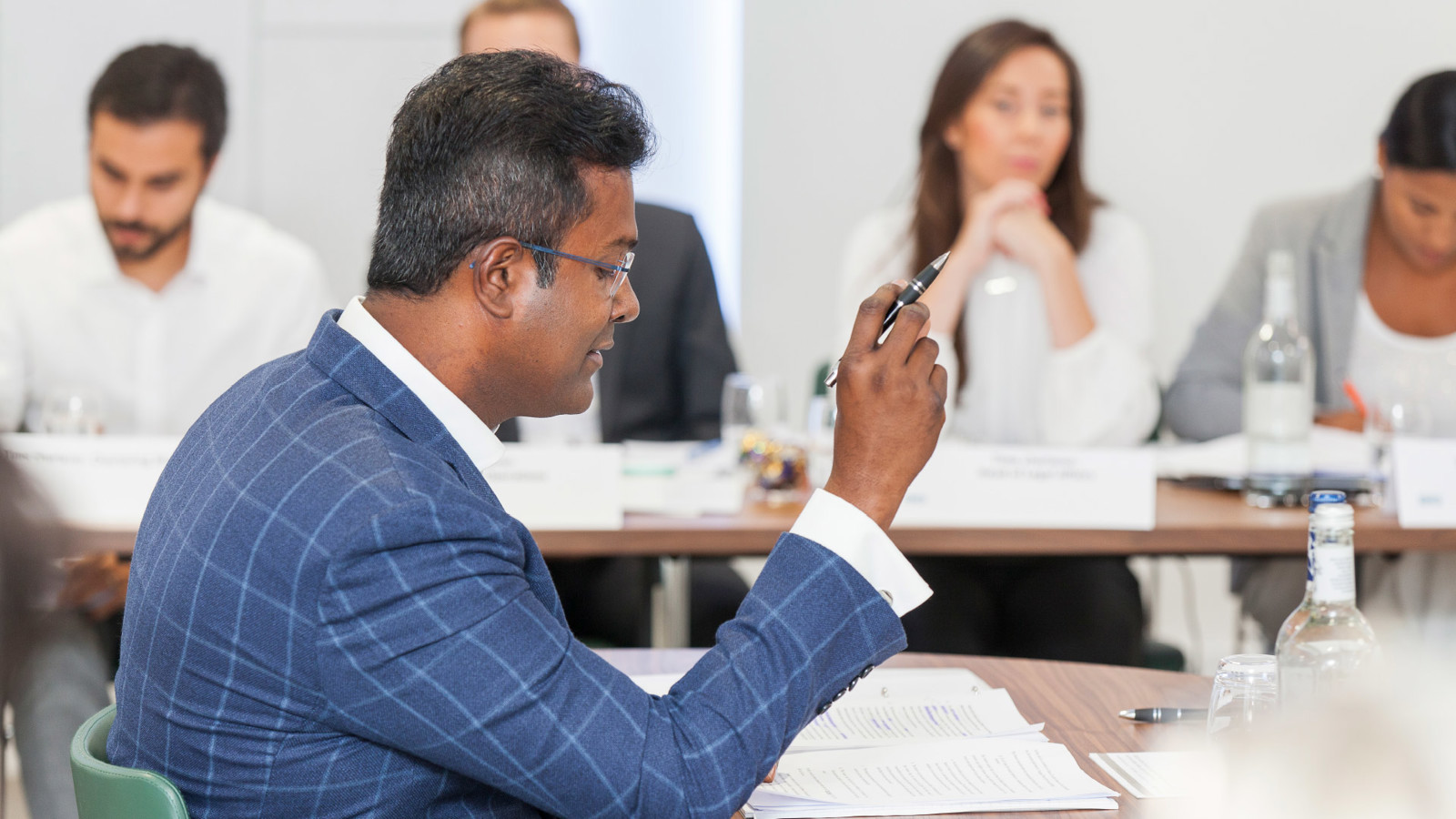 Man in suit sitting at a table reading papers and holding a pen up with a row of blurred people watching him in the background