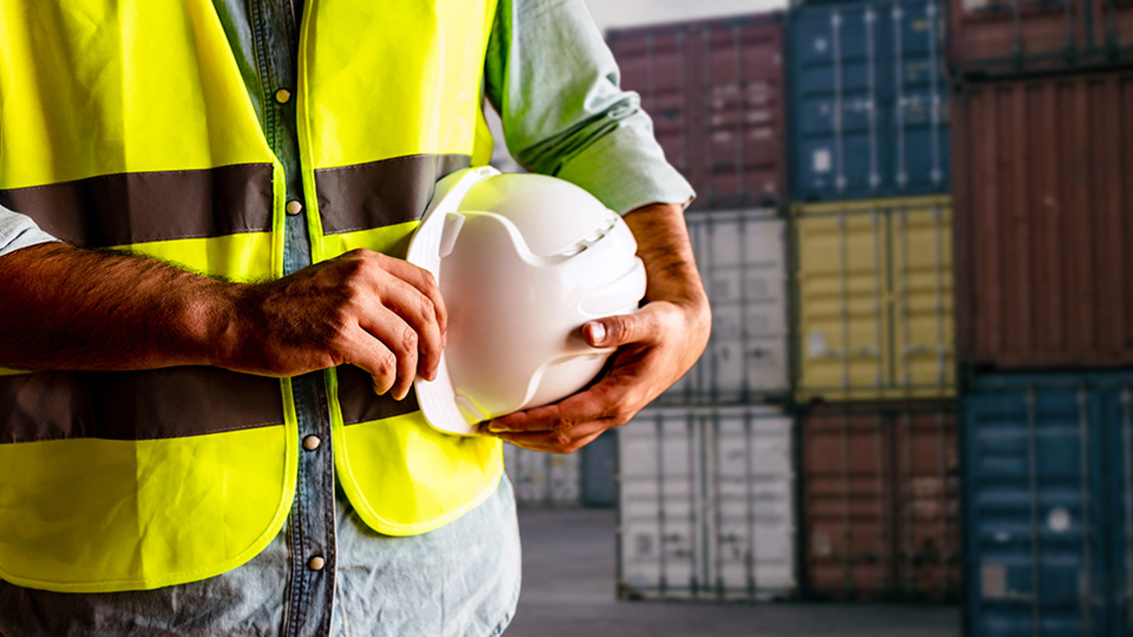 A dockworker holding a safety hat and wearing a yellow hi-vis vest stands in front of shipping containers
